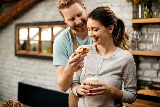 Happy couple having fun while eating in the kitchen. Man is feeding girlfriend with a sandwich.  - Foto, Imagem