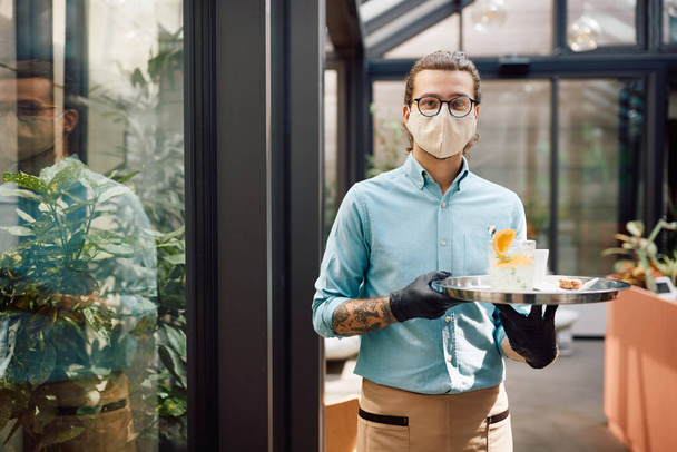 Waiter with protective face mask serving drinks while working at cafe during coronavirus pandemic and looking at camera. - Фото, изображение