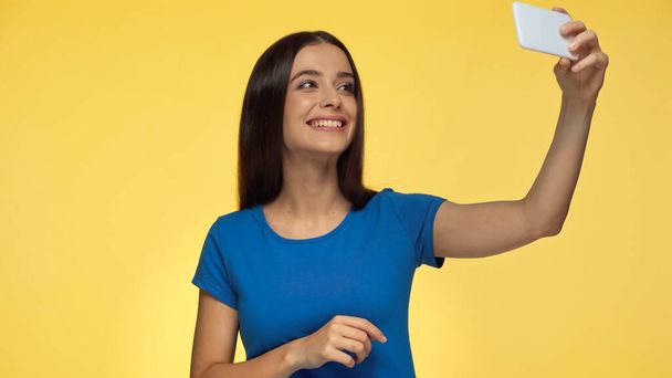 young brunette woman in blue t-shirt smiling while taking selfie isolated on yellow - Fotó, kép