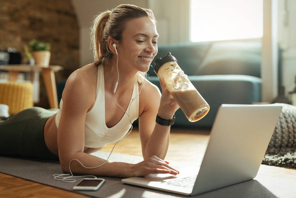 Young happy sportswoman drinking smoothie while relaxing on the floor and surfing the net on laptop.  - Photo, image