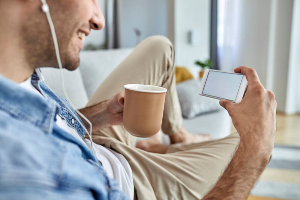 Close-up of  man drinking coffee while using smart phone and relaxing in the living room. Focus is on blank device screen. Copy space.  - Fotografie, Obrázek