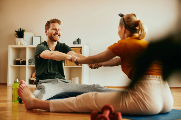 Happy athletic couple holding hands and stretching during home workout. Focus is on man.  - Photo, Image