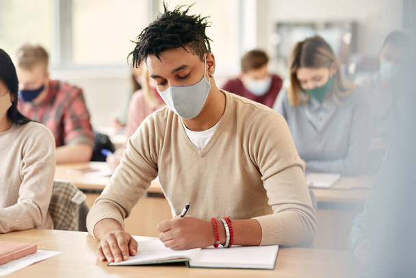 African American student writing in notebook while wearing face mask and attending lecture at the university during coronavirus pandemic. - Фото, изображение