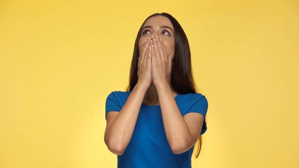 emotiva giovane donna in t-shirt blu che copre la bocca e alza lo sguardo isolato sul giallo - Foto, immagini