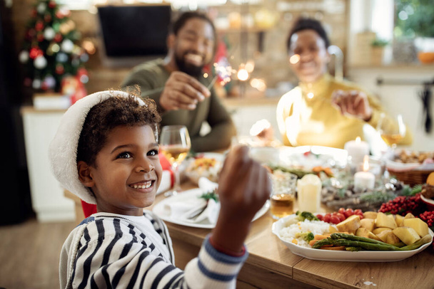 Happy African American boy and his parents using sparklers and having fun during Christmas lunch in dining room.  - 写真・画像