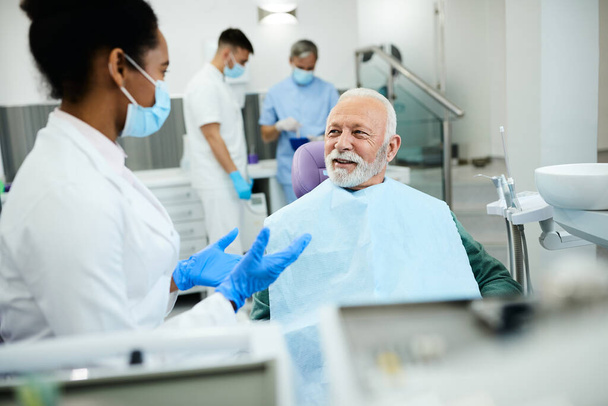 Black female dentist communicating with mature man during teeth exam at dentist's office. Focus is on man.  - Foto, immagini