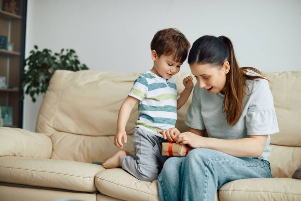 Happy mother and son untying ribbon while opening present together at home. Focus is on boy. - Foto, afbeelding