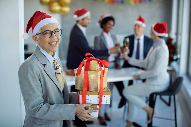 Boldog, érett üzletasszony ajándékdobozokkal, aki eljön az irodai karácsonyi partira és kamerába néz. A kollégái ünnepelnek a háttérben..  - Fotó, kép