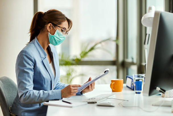 Молода бізнес-леді носить захисну маску для обличчя, проходячи доповіді та працюючи в офісі
.  - Фото, зображення