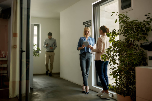 Ευτυχισμένες επιχειρηματίες που επικοινωνούν ενώ στέκονται στο διάδρομο ενός κτιρίου γραφείων. Εστίαση στην επιχειρηματία που κατέχει ψηφιακή ταμπλέτα. - Φωτογραφία, εικόνα