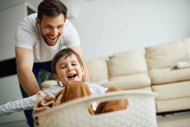Щасливий батько штовхає свого маленького сина в кошик для білизни, граючи разом вдома. Фокус на хлопчику
.  - Фото, зображення