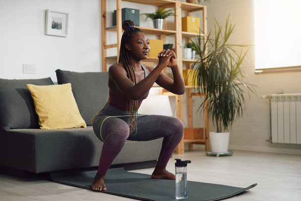 Щаслива чорна атлетична жінка робить присідання з групою опору навколо її ніг вдома
. - Фото, зображення