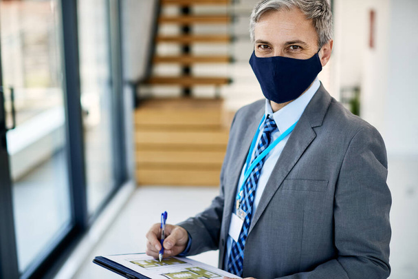 Усміхнений бізнесмен аналізує плани нерухомості і носить захисну маску для обличчя через пандемію коронавірусу
.  - Фото, зображення