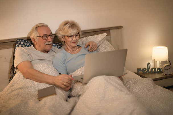 Mosolygó idős pár pihentető a hálószobában, és szörfözés az interneten a laptop.  - Fotó, kép