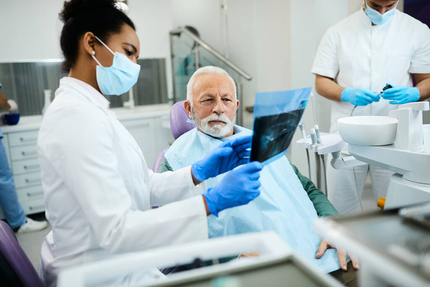 African American dentist showing to senior man his orthopantomogram during dental appointment. - Φωτογραφία, εικόνα