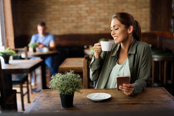 Νεαρή ευτυχισμένη γυναίκα πίνει καφέ ενώ χρησιμοποιεί το κινητό τηλέφωνο και χαλαρώνει σε ένα καφέ.  - Φωτογραφία, εικόνα