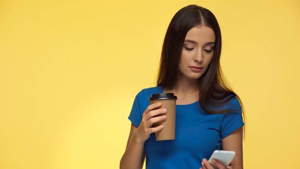 mladá brunetka žena v modrém tričku pomocí smartphonu a držení kávy jít izolované na žluté - Fotografie, Obrázek