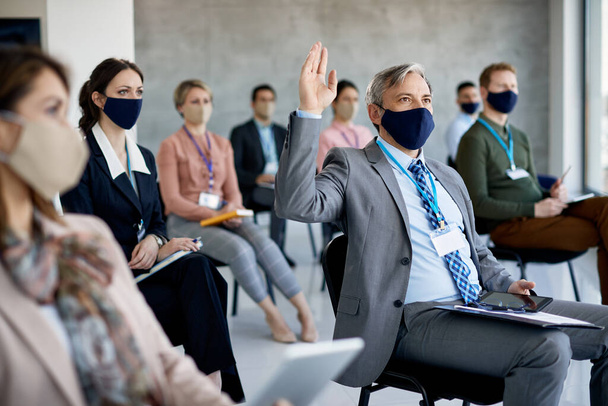 Μεγάλη ομάδα επιχειρηματιών που παρακολουθούν επιχειρηματικό σεμινάριο και φορούν προστατευτικές μάσκες προσώπου λόγω πανδημίας COVID-19. Εστίαση στον επιχειρηματία με σηκωμένο χέρι..  - Φωτογραφία, εικόνα