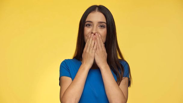 young and excited woman in blue t-shirt covering mouth while looking at camera isolated on yellow - Фото, изображение