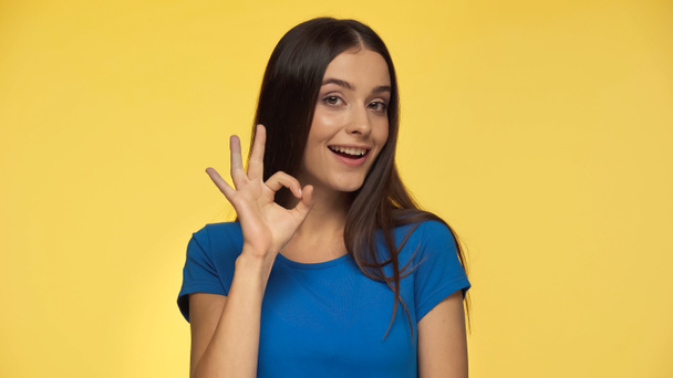 young and cheerful woman in blue t-shirt showing ok sign isolated on yellow - Photo, Image