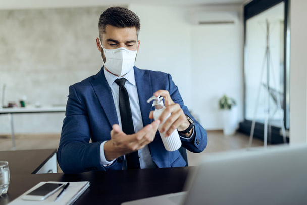 Businessman with face mask disinfecting hands while working at office desk.  - Φωτογραφία, εικόνα