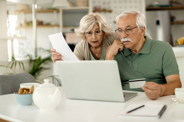 Worried mature couple using laptop and credit card while checking their online bank account and analyzing their finances at home.  - 写真・画像