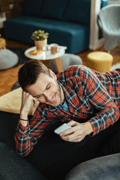 Vysoký úhel pohledu na mladého šťastného muže pomocí mobilního telefonu a textové zprávy při ležení na pohovce.  - Fotografie, Obrázek