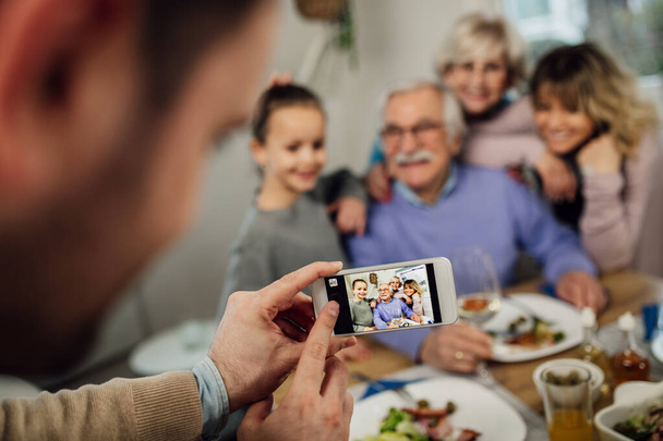 Close-up of man using smart phone while photographing his happy extended family in dining room.  - Фото, изображение