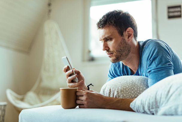 Young man lying down in bed and drinking coffee while text messaging on mobile phone.  - Foto, Imagem
