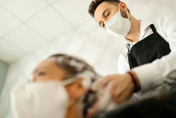 Низький кут зору перукаря з захисною маскою для обличчя, що миє волосся клієнта в салоні під час епідемії коронавірусу
.  - Фото, зображення
