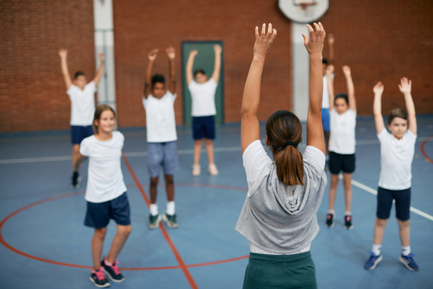 Задний вид учителя физкультуры и группы учащихся начальных классов во время занятий в школьном спортзале.  - Фото, изображение