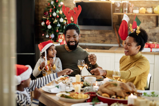 Joyful African American family using sparklers while having lunch and celebrating Christmas at dining table.  - Фото, изображение