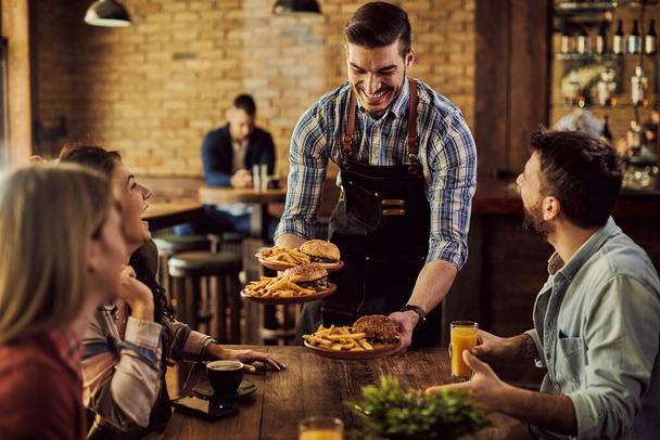 Group of happy friends having fun while waiter is serving them food in a pub. Focus is on waiter.  - Zdjęcie, obraz