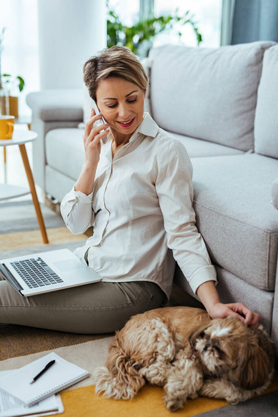 Smiling freelance worker with a dog communicating on cell phone while working on laptop at home.  - 写真・画像