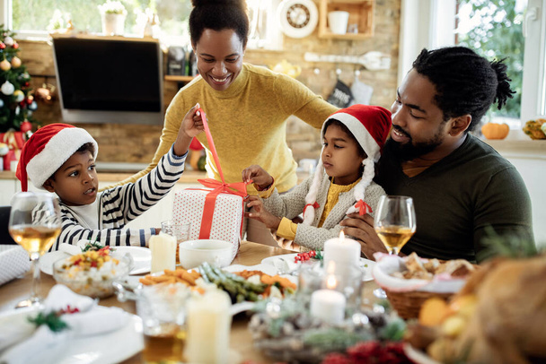 Szczęśliwi czarni rodzice z dziećmi otwierając prezenty świąteczne podczas rodzinnego lunchu w jadalni. - Zdjęcie, obraz