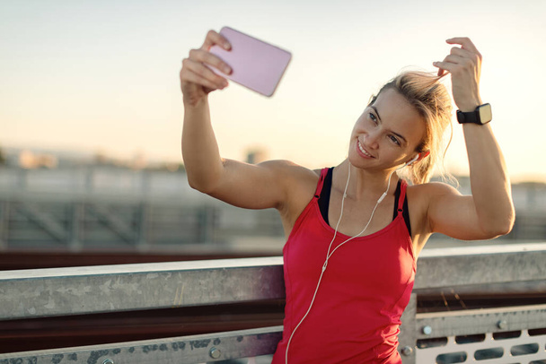 Smiling athletic woman using mobile phone and taking selfie while working out outdoors.  - Foto, imagen