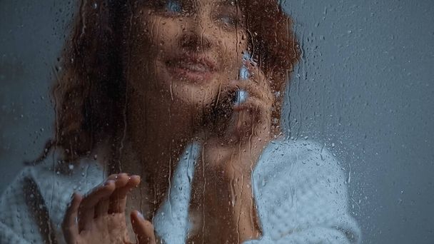 cheerful and curly woman talking on smartphone behind window glass with raindrops - Photo, image