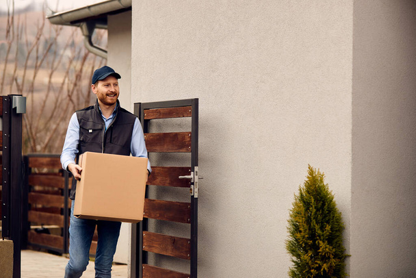 Smiling male courier carrying cardboard box while making home delivery to his customer.  - Foto, Bild