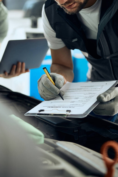 Close-up van auto monteur schrijven van gegevens tijdens het onderzoek van motorproblemen met zijn collega in de reparatiewerkplaats.  - Foto, afbeelding