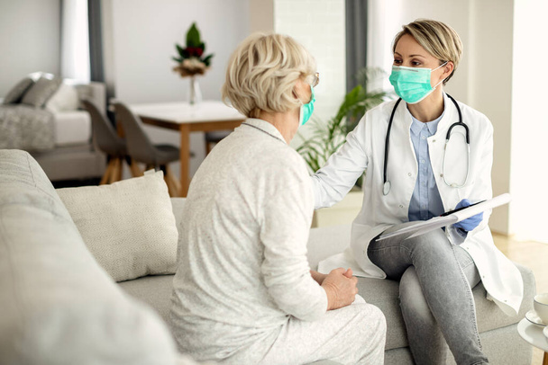 Female doctor and senior woman talking during home visit and wearing protective face masks due to coronavirus pandemic.  - Foto, Bild