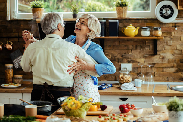 Carefree mature woman dancing with her husband while preparing food in the kitchen.  - Φωτογραφία, εικόνα