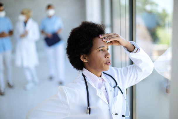 Black female doctor feeling worried and thinking of something while looking through the window in the hospital.  - Foto, Bild
