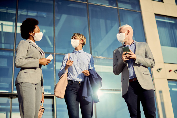 Low angle view of business coworkers with protective face masks talking while standing in front of their office building.  - Foto, Imagem
