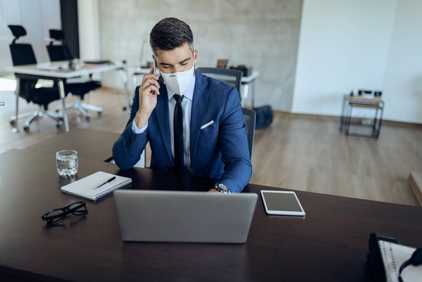Businessman wearing protective face mask while working on laptop and communicating over cell phone in the office. - Foto, Imagen
