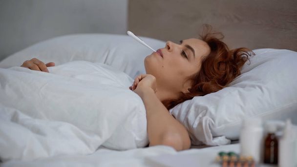 redhead and sick woman measuring temperature with electronic thermometer and lying on bed - Zdjęcie, obraz