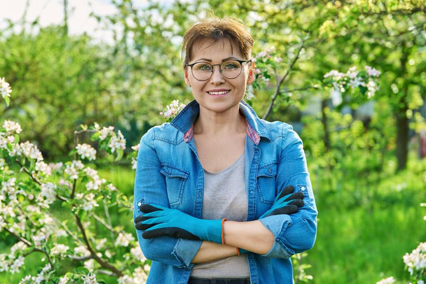 Portrait of beautiful middle aged woman in blooming spring garden. Smiling confident female in gardening gloves with crossed arms looking at camera. Springtime, nature, orchard, 40s people concept - Фото, изображение