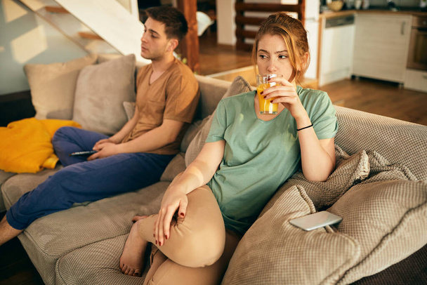 Молода жінка п'є апельсиновий сік, розслабляючись на дивані і дивлячись телевізор зі своїм хлопцем у вітальні
.  - Фото, зображення
