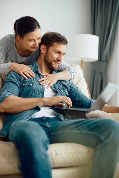 Smiling woman embracing her husband who is using laptop while relaxing on the sofa at home.  - Valokuva, kuva