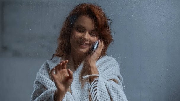 smiling and curly woman talking on smartphone behind window glass with raindrops - Foto, Imagen