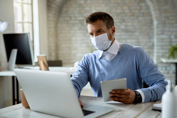 Бізнесмен в захисній масці для обличчя під час роботи на комп'ютері та використання цифрового планшета в офісі
.  - Фото, зображення